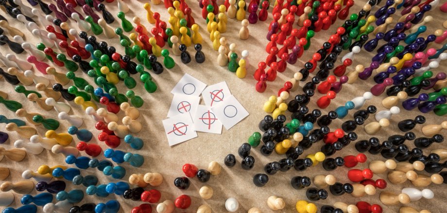 Vogelperspektive auf bunte Holzspielfiguren, welche um Blätter mit angekreuzten Kreisen stehen. Symbolbild für Wahlen